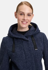 Granatowy, strukturalny płaszcz wełniany z kapturem, z czarnymi sznurkami i akcentami z logo. Kołnierz jest wyściełany i sięga do górnej części klatki piersiowej.