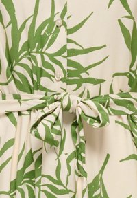 Crèmekleurige stof met groen tropisch bladprint, drie knopen aan de voorkant en een stoffen ceintuur gestrikt in de taille.