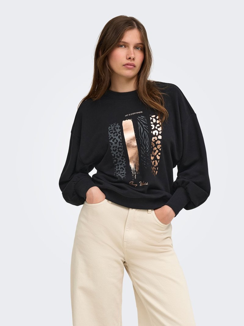Jeune femme portant un sweat-shirt noir avec des rayures brillantes à motif animalier et un pantalon large beige, debout les mains dans les poches.