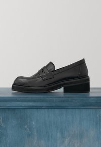 Mocassin noir en cuir avec un talon bas et épais reposant sur une surface en bois bleu sur un fond clair uni.