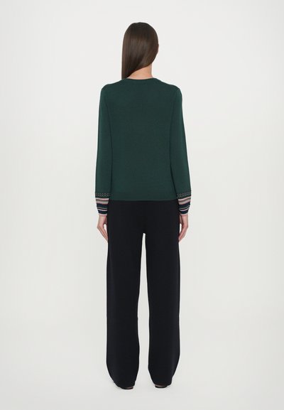 Pulover verde închis cu mâneci lungi, având accente cu dungi la manșete; asortat cu pantaloni negri, largi. Textură netedă.