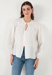 Bjela, bluzica dugih rukava s okruglim izrezom, detaljem kravate, kopčanjem sprijeda i peplum krojem. Izrađena od laganog materijala s teksturiranim završetkom.
