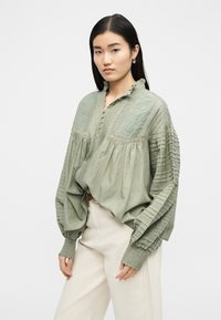 Γυναίκα που φοράει μπλούζα σε χρώμα φασκόμηλου με δαντέλα και πιετάκια, φουσκωτά μανίκια και λευκό ψηλόμεσο παντελόνι, στέκεται μπροστά σε λευκό φόντο.