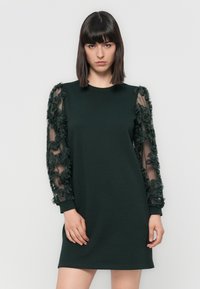 Woman with straight dark hair wearing a black knee-length dress with sheer floral-embellished long sleeves, posing against a plain background.