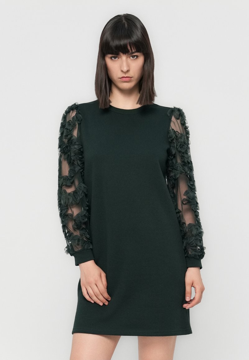 Frau mit glattem, dunklem Haar, die ein schwarzes knielanges Kleid mit durchsichtigen, floral verzierten langen Ärmeln trägt, posiert vor einem schlichten Hintergrund.