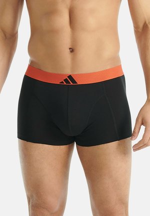 Torse masculin portant un boxer noir avec une ceinture orange arborant le logo Adidas, se tenant devant un fond uni.