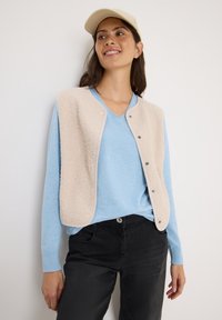 Beige fleece vest met drukknoopjes over een lichtblauwe V-hals sweater, gecombineerd met zwarte broek. Model draagt een beige pet en staat tegen een effen achtergrond.