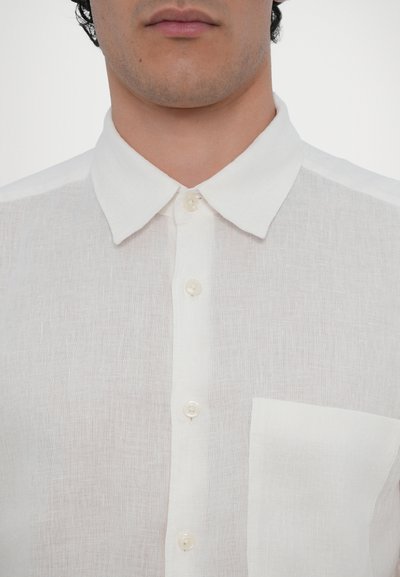 Camisa de linho, branca, com gola colarinho, com abotoamento à frente e um único bolso no peito. Tecido texturizado com uma aparência ligeiramente translúcida.