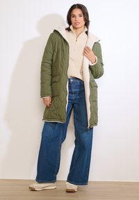 Žena v otvorenom zelenom prešívanom kabáte s fleece podšívkou, béžovom svetri, širokých modrých džínsoch a béžových teniskách stojaca vo vnútri.