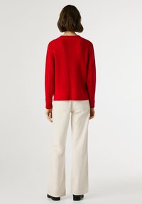 Kvinna som står med ryggen mot kameran, iklädd en röd stickad tröja, off-white vidbyxor och svarta skor mot en neutral vit bakgrund.