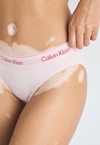 A Calvin Klein rózsaszín fehérnemű sima anyaggal, kontrasztos piros logós derékpánttal és klasszikus bugyi szabással rendelkezik, kiemelve a derekat.