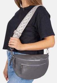 Sac bandoulière en cuir gris avec surface texturée, comprenant deux poches zippées et une sangle réglable à motif. Forme arrondie et accents subtils.
