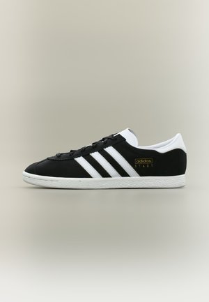Baskets Adidas noires avec bandes et semelle blanches, logo doré "adidas STADT", design bas et languette de talon blanche sur fond neutre.