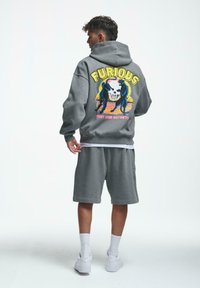Grå hettegenser med grafikk som viser et skalle og bjørner, med teksten "FURIOUS", paret med matchende grå shorts og hvite sneakers.