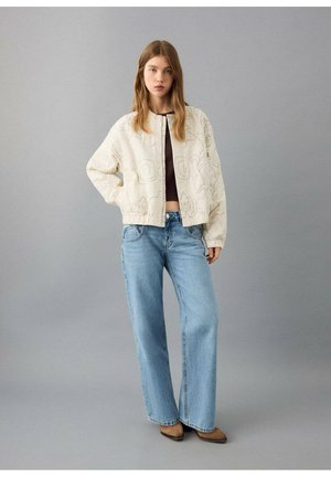 Bomber jacket crema con ricami floreali, abbinata a jeans a gamba larga color azzurro chiaro. La modella posa su uno sfondo grigio neutro.