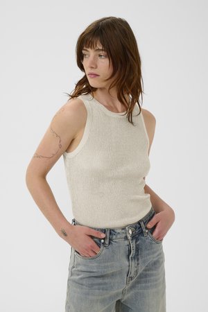 Jeune femme aux cheveux bruns porte un haut beige sans manches et un jean bleu clair, les mains dans les poches, regardant sur le côté sur un fond uni.