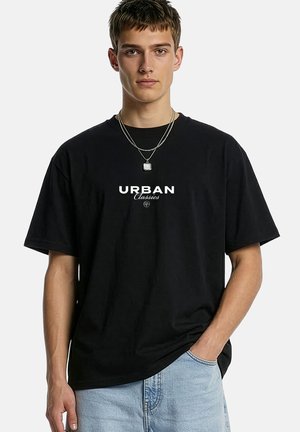 Jeune homme portant un t-shirt noir "Urban Classics", un jean bleu clair et des colliers argentés en couches, debout avec une main dans la poche.