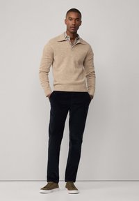 Beige knitted sweater with a collar, layered over a checked shirt. Navy corduroy pants and olive green sneakers complete the outfit.