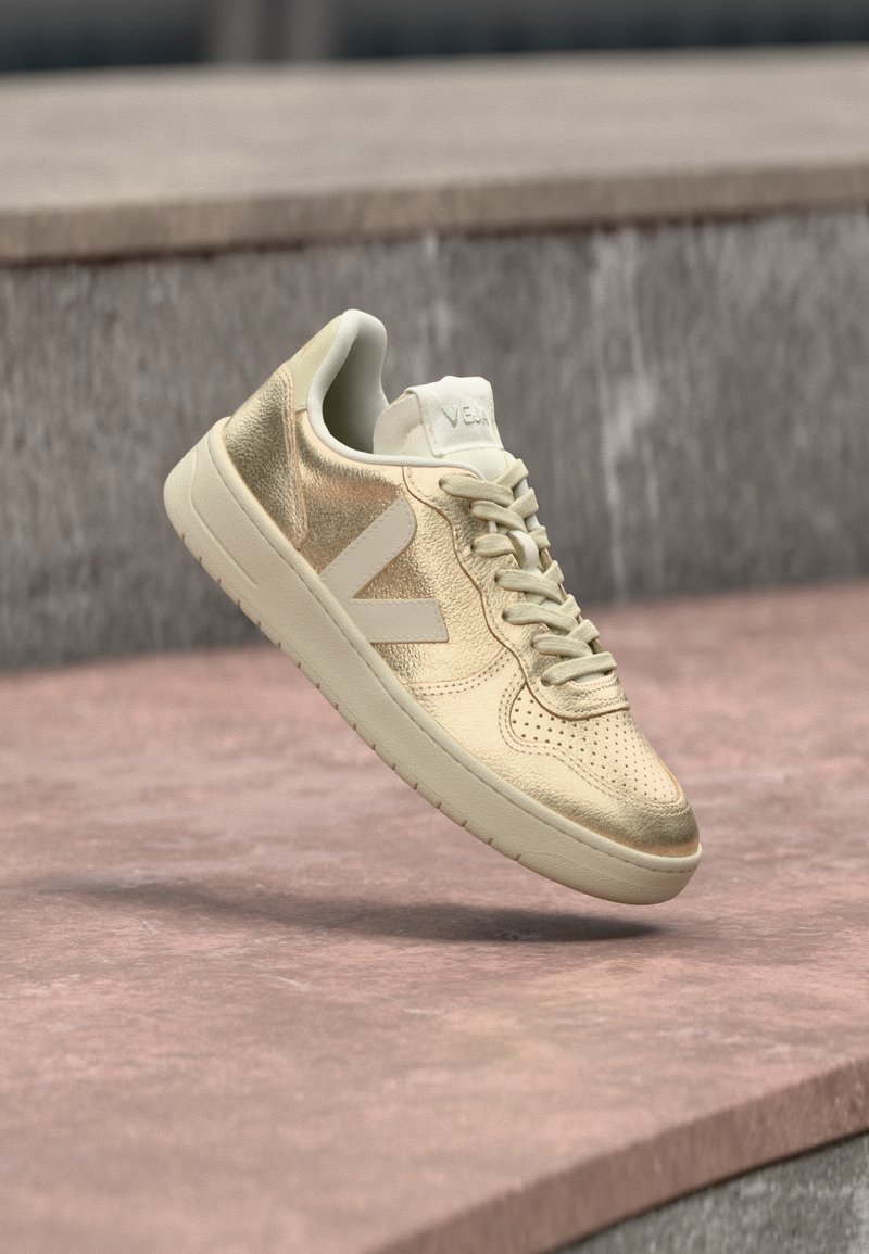 Χρυσά δερμάτινα αθλητικά παπούτσια με διάτρητο μπροστινό μέρος, επίπεδα εκρού κορδόνια και ουδέτερη λαστιχένια σόλα. Διαθέτουν λογότυπο στη γλώσσα και στην πλευρά.