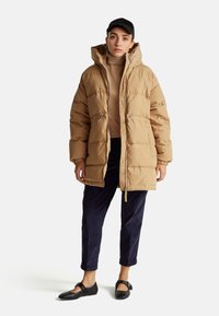 Manteau matelassé beige avec capuche et fermetures à bascule, porté sur un col roulé crème. Associé à un pantalon en velours côtelé foncé et des ballerines noires.