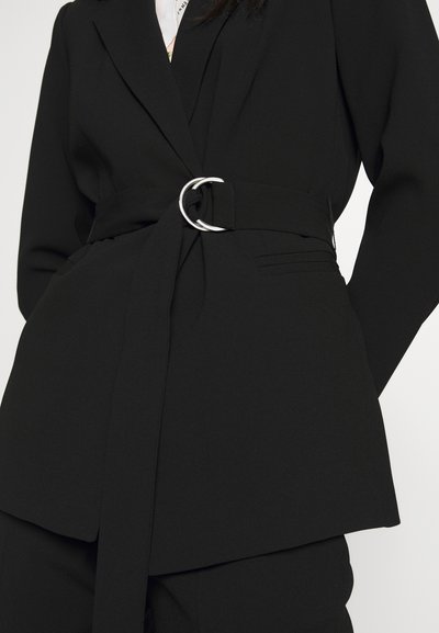 Blazer nero con cintura e anello d'argento a cerchio. Presenta spalle strutturate, vestibilità sartoriale e tasche su entrambi i lati. Tessuto testurizzato.