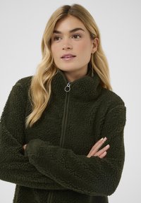 Oliwkowa zielona kurtka polarowa z wysokim kołnierzem, zamkiem błyskawicznym i teksturowanym, przyjemnym w dotyku materiałem. Posiada długie rękawy i luźny krój.