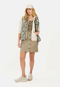 Gevoerde vest met groen en crème dierenprint, gecombineerd met een beige top en een knopenrok in khaki. Witte sneakers met lichtbruine accenten.
