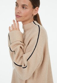 Beige knitted sweater with a high collar, ribbed texture, and black stripe detail along the shoulder and sleeve edges.