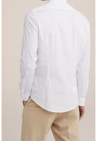 White long-sleeve shirt with a curved hem, buttoned cuffs, and a smooth texture. Worn with beige trousers, showcasing a clean, classic design.