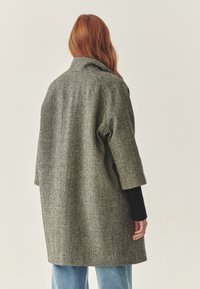 Cappotto grigio oversize con un tessuto texture, maniche ampie e colletto alto, indossato sopra un maglione nero e pantaloni azzurro chiaro.