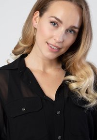 Zwarte, doorzichtige knoopshirt met twee borstzakken. Model heeft golvend, blond haar en draagt een delicate gouden ketting.