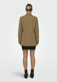 Hnědý přizpůsobený blazer se strukturovaným siluetu, s jedním zadním rozventem a špičatým klopou, v kombinaci s černou mini sukni a kotníkovými botami.
