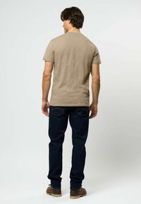 Vyras, vilkintis smėlio spalvos trumpomis rankovėmis marškinėlį, tamsiai mėlynas džinsus ir rudas batus, stovintis nugara į žiūrovą prieš paprastą šviesų foną.