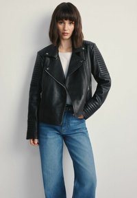 Czarna skórzana kurtka motocyklowa z pikowanymi akcentami na ramionach, zapinana na zamek z szerokimi klapami. Połączona z jasnoniebieskimi spodniami typu wide-leg.