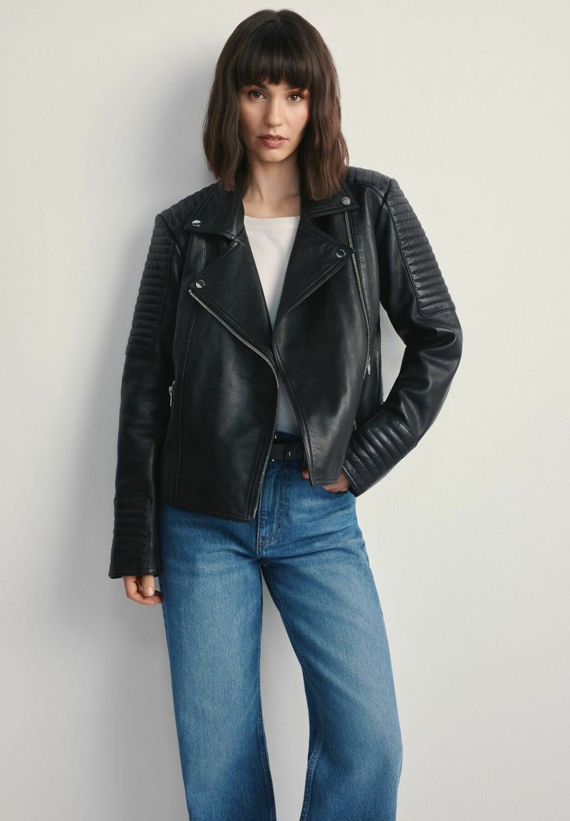 Czarna skórzana kurtka motocyklowa z pikowanymi akcentami na ramionach, zapinana na zamek z szerokimi klapami. Połączona z jasnoniebieskimi spodniami typu wide-leg.