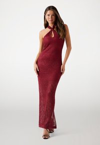 Abito lungo in pizzo bordeaux con collo all'americana e dettaglio a chiave. Silhouette aderente, trama intricata e spacco laterale. Abbinato a tacchi neri con cinturini.