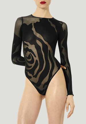 Frau trägt einen schwarzen transparenten Body mit abstrakten wirbelnden Mustern und langen Ärmeln, steht mit einem Arm hinter dem Rücken angewinkelt.