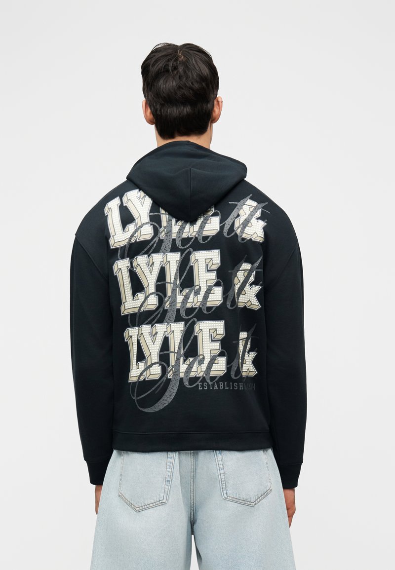 Άνδρας που φοράει μαύρο φούτερ με κουκούλα με μεγάλα λευκά γράμματα "LYLE &" επαναλαμβανόμενα τρεις φορές στην πλάτη, σε συνδυασμό με ανοιχτό μπλε τζιν.