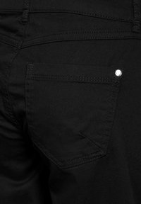 Schwarze Denim-Jeans mit einer einzigen Gesäßtasche, sichtbaren Nähten und einem silbernen Metallnieten-Detail. Glatte Textur, eng anliegendes Design.