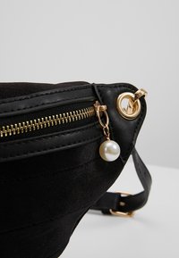 Sac en tissu noir avec fermeture éclair dorée, orné d'un accent de perle sur une boucle dorée et une fine bandoulière noire. Texture lisse, forme triangulaire.