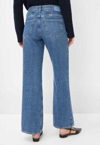 Lange blauwe denim jeans met een gemiddelde wassing, achterzakken en een hoge taille. Gestyld met een zwarte top en zwarte sleehakken.