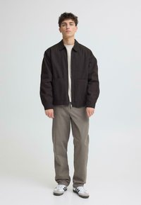 Veste noire zippée à coupe décontractée avec deux poches avant, portée sur une chemise crème et associée à un pantalon gris et des baskets blanches.