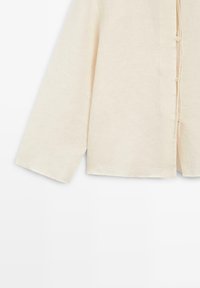 Beżowa koszula z długim rękawem na guziki, luźnego kroju z widocznym troczkiem w pobliżu guzików na białym tle.