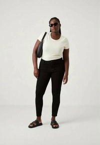 Top blanc à rayures, manches courtes, jean skinny noir taille haute, sandales noires et sac à bandoulière noir. Le mannequin se tient contre un fond clair.