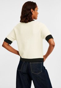 Person sedd bakifrån som bär en vit kortärmad tröja med svarta mudd och fåll, i kombination med mörkblå jeans.