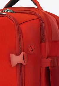 Mochila de tela roja con una superficie texturizada, múltiples cremalleras, un asa resistente y una insignia de logo redonda con un patrón de cruces.