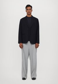 Zwarte blazer met twee knopen, gecombineerd met lichtgrijze geplooide broek en zwarte schoenen. Eenvoudig ontwerp met een nauwsluitende pasvorm.