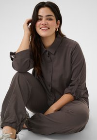 Chemise marron à boutons avec manches retroussées, portée avec un pantalon décontracté assorti. Le tissu semble lisse, avec un design décontracté et des poignets élastiques.