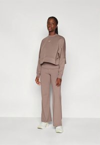 Barna rövid felső fehér logóval és hosszú ujjú kialakítással, melyet hozzáillő barna bővülő szárú nadrággal viselnek. A modell fehér sportcipőt hord.