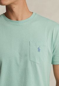 Ljusgrön kortärmad T-shirt i bomull, med en bröstficka till vänster som har en liten blå broderad logotyp. Mjuk textur.